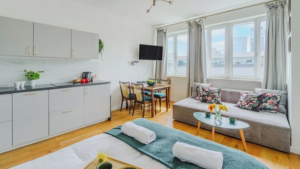 A modern hotel room with a kitchenette, a dining area, a sofa with floral cushions, and a large window offering a city view. - ShortStayPoland Plac Mirowski (Warsaw)