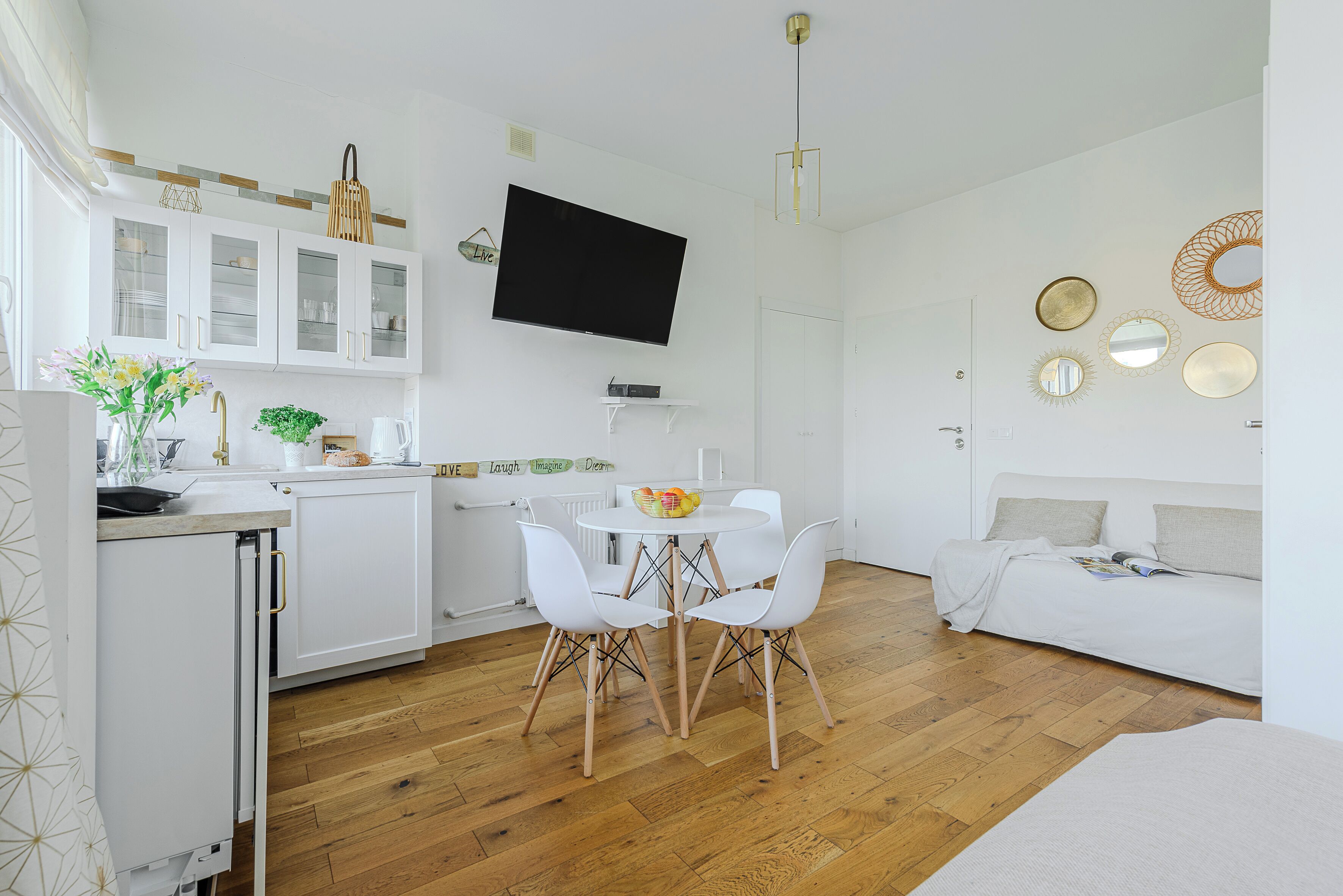 A modern kitchen with white cabinets, a dining area with a round table and chairs, and a living area with a sofa and decorative mirrors.