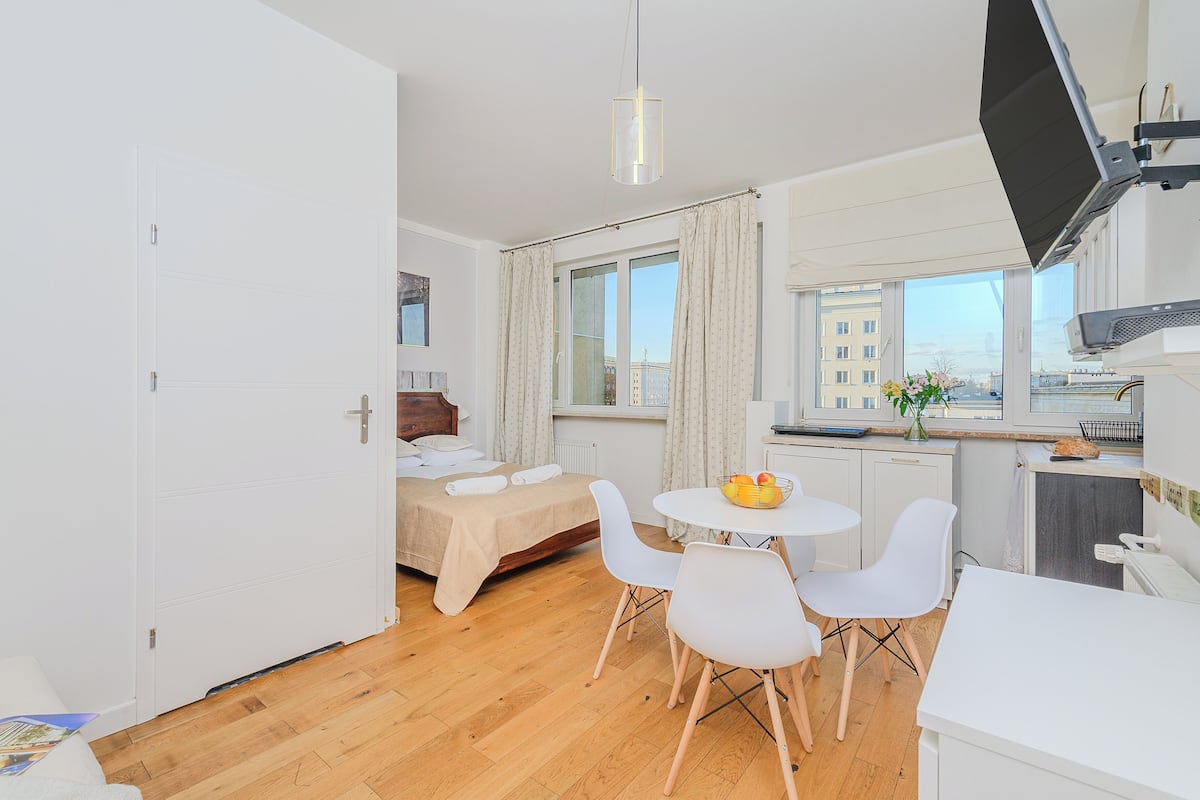Ein kompaktes Hotelzimmer mit Bett, Essbereich mit Tisch und Stühlen, einer Küchenzeile und einem großen Fenster mit Blick auf die Stadt.