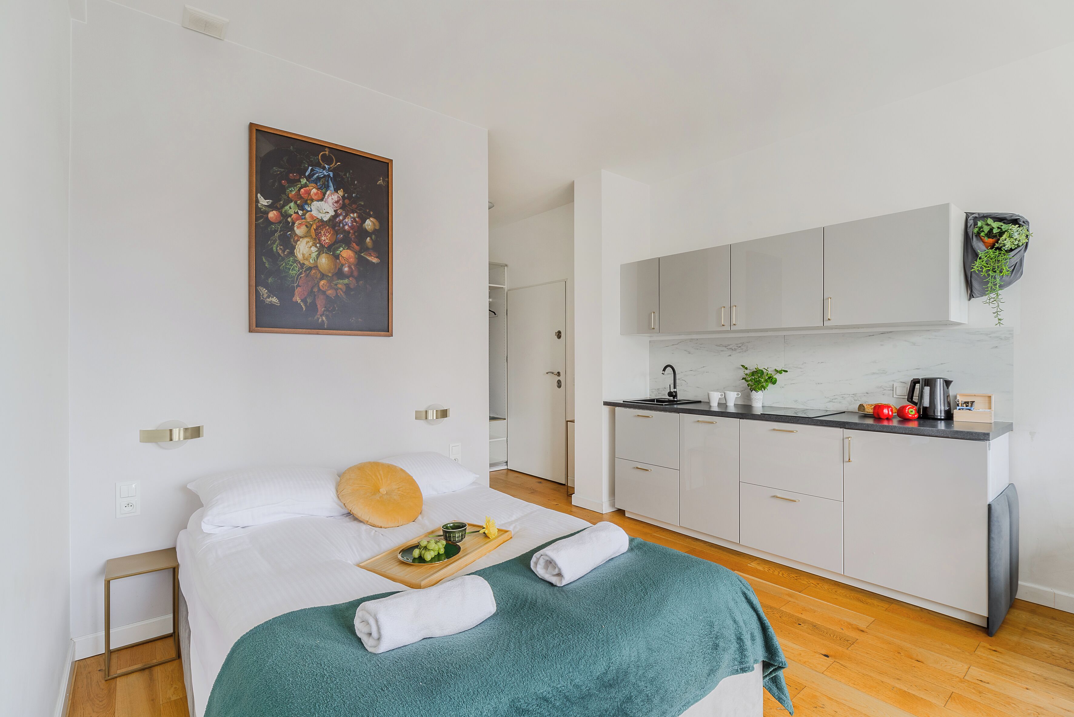 Une chambre d’hôtel moderne avec un lit, une petite table de chevet, un plaid vert, une serviette, un panier de fruits et un tableau au mur.