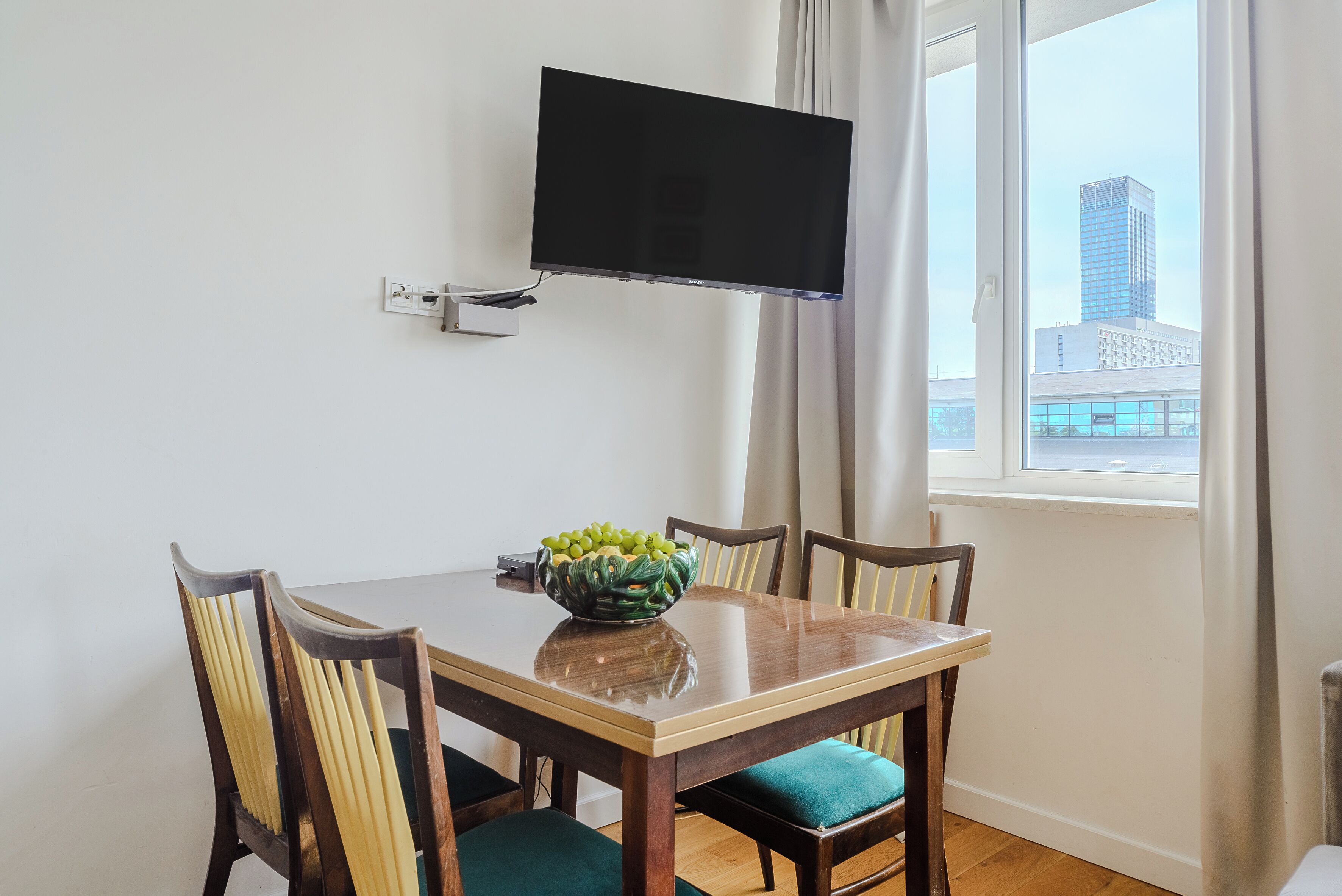 A dining area with a table, chairs, a TV mounted on the wall, and a window with a city view.