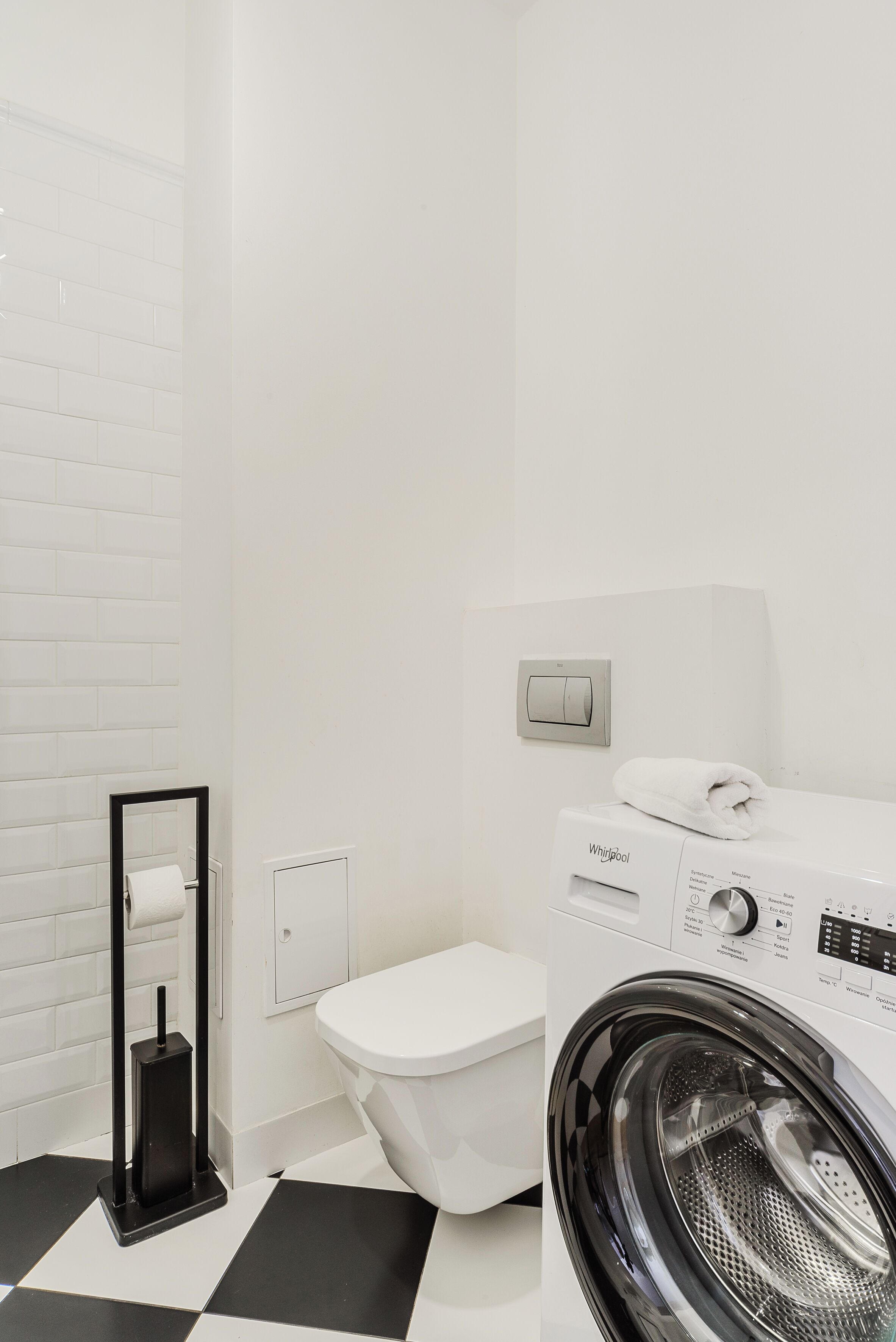 Un bagno compatto con un water bianco, un pavimento a scacchi bianconero, una parete in mattoni bianchi, un porta scopettone nero e una lavatrice Whirlpool bianca.