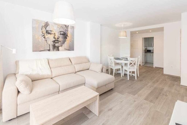 Una sala de estar moderna con un sofá seccional color beige, una mesa de centro de madera y una zona de comedor con sillas blancas y una mesa.