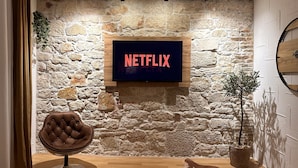 Una habitación con pared de piedra, un televisor montado mostrando 'NETFLIX', un sillón de cuero y una mesa de madera.
