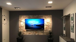 A living room with a stone wall, a flat-screen TV, two potted plants, and a console table with framed pictures. - Apartment Porta Aurea (Pula)