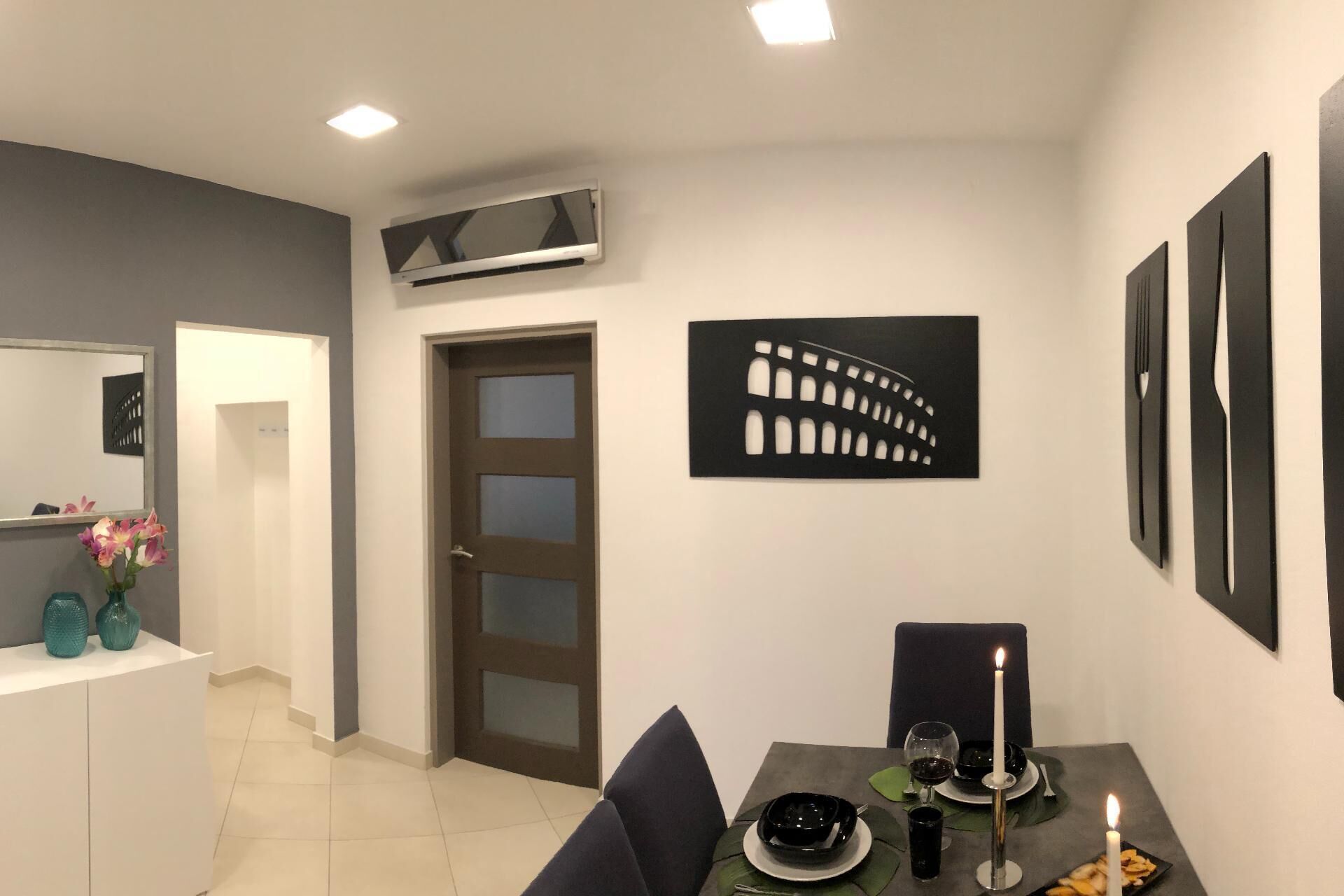 A modern dining area with a table set for two, a black and white wall art piece, and a ceiling-mounted air conditioning unit.