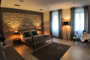 Ein Schlafzimmer mit Steinwand, einem Bett mit Metallgestell, zwei Nachttischlampen, einer Stehlampe, einem Sessel und einem kleinen Tisch mit einer Vase.