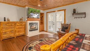 A log cabin interior with a fireplace, wooden furniture, and a large window with a view of a waterfall. - Secluded Smoky Mountain Cabin with Private Waterfall, Creek, & Luxury Amenities! (Sevierville)