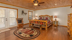 A log cabin bedroom with a wooden ceiling, a bed with a plaid blanket, a chair, a small table with a lamp, and a rug with animal prints. - Secluded Smoky Mountain Cabin with Private Waterfall, Creek, & Luxury Amenities! (Sevierville)