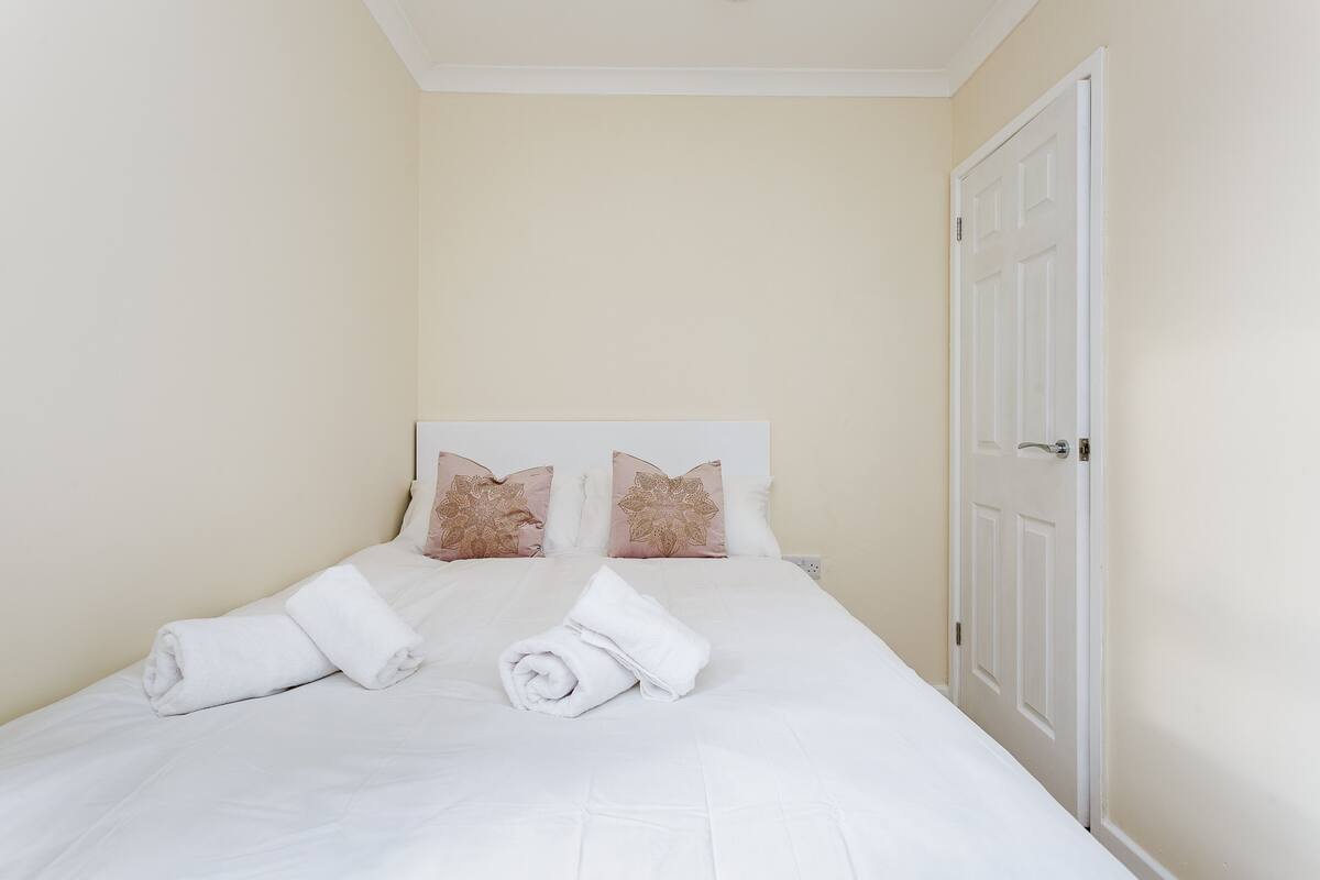 A neatly made bed with white linens and two decorative pillows, a white door, and cream-colored walls.