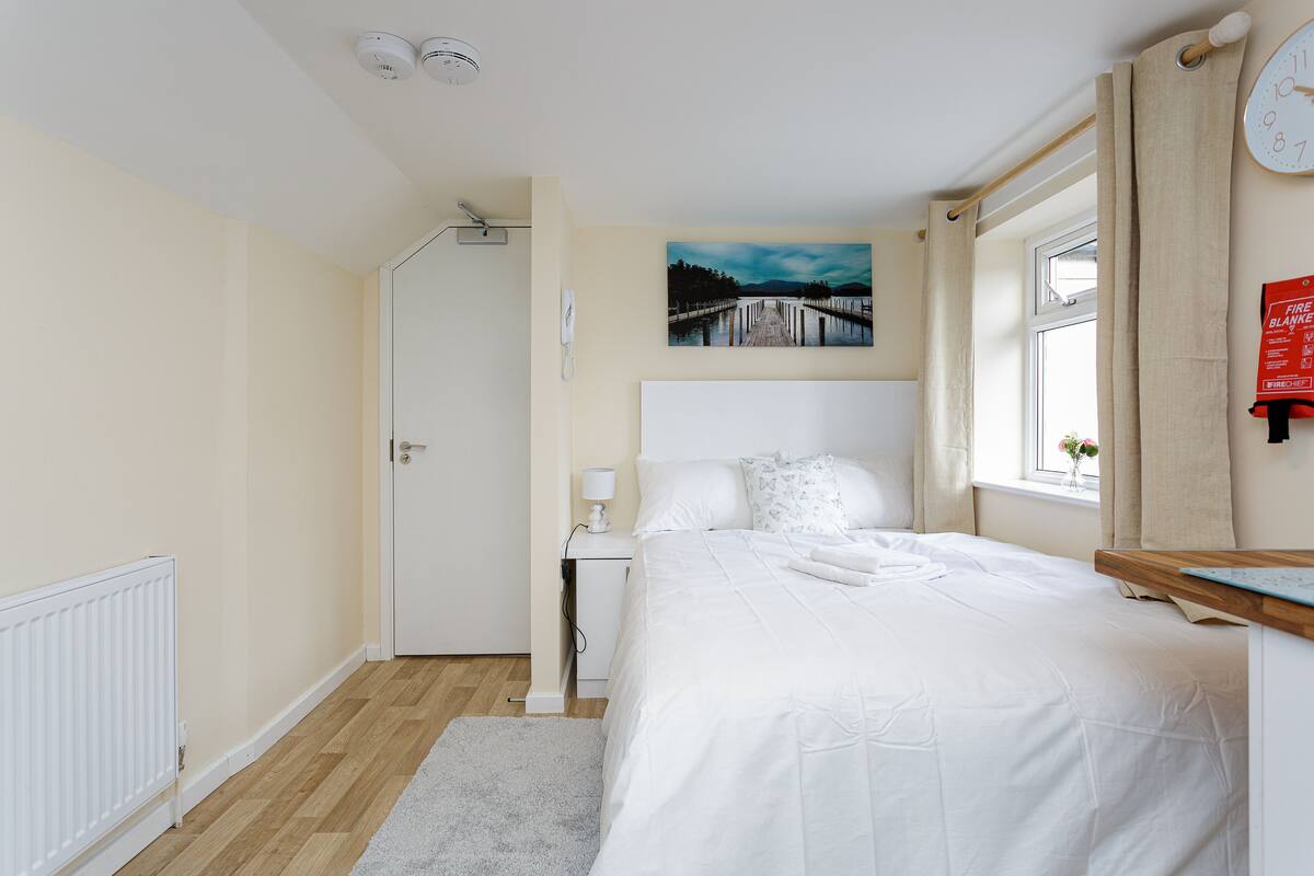 A bedroom with a bed, a bedside table with a lamp, a painting on the wall, a window with curtains, and a fire blanket on the wall.