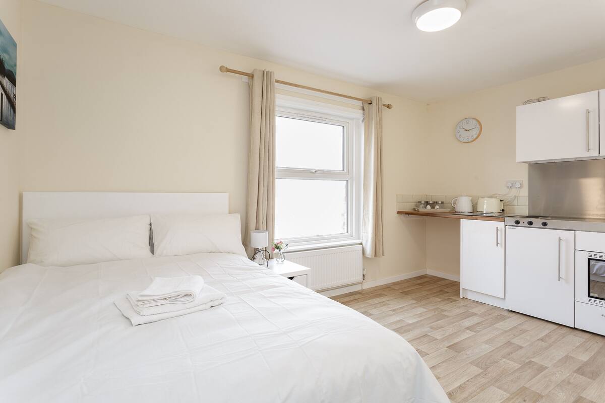 A bedroom with a bed, a nightstand, a window with curtains, a wall clock, and a kitchenette with cabinets and a stove.