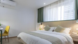 A hotel room with a large bed, a desk with a chair, a bedside table with a lamp, and a window with curtains.