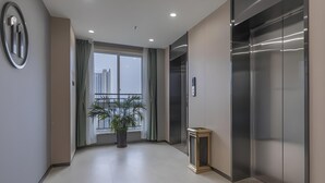 A modern elevator with a metallic finish, a potted plant in the hallway, and a view of a cityscape through a large window.