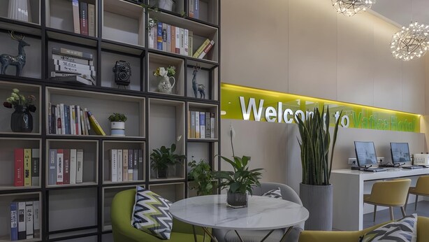 A modern reception area with a 'Welcome to' sign, a desk with two monitors, a round table with a plant, and a bookshelf filled with books and decorative items.