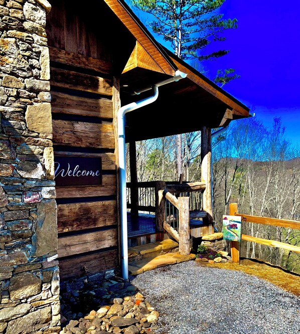 A rustic wooden cabin with a stone wall, a wooden porch, and a 'Welcome' sign. - Cedar Cliff Cabin at Rivers Edge Rentals. Luxurious reclaimed 125 year old cabin (Waynesville)