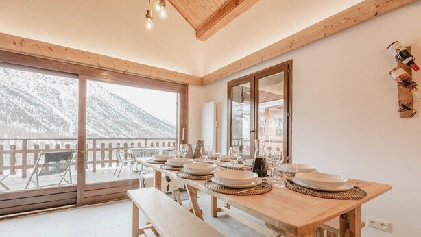 Zona pranzo con tavolo in legno apparecchiato per quattro persone, ampie finestre e vista sulle montagne innevate.