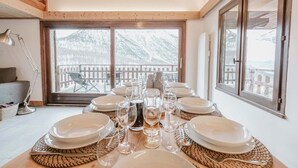 Tavola imbandita con piatti, bicchieri e una caraffa, vista su montagne innevate attraverso ampie finestre.