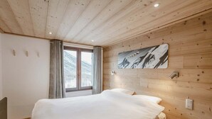 Una camera da letto con soffitto in legno, un letto con copripiumino bianco, una finestra con tende e un'opera d'arte a parete raffigurante montagne.