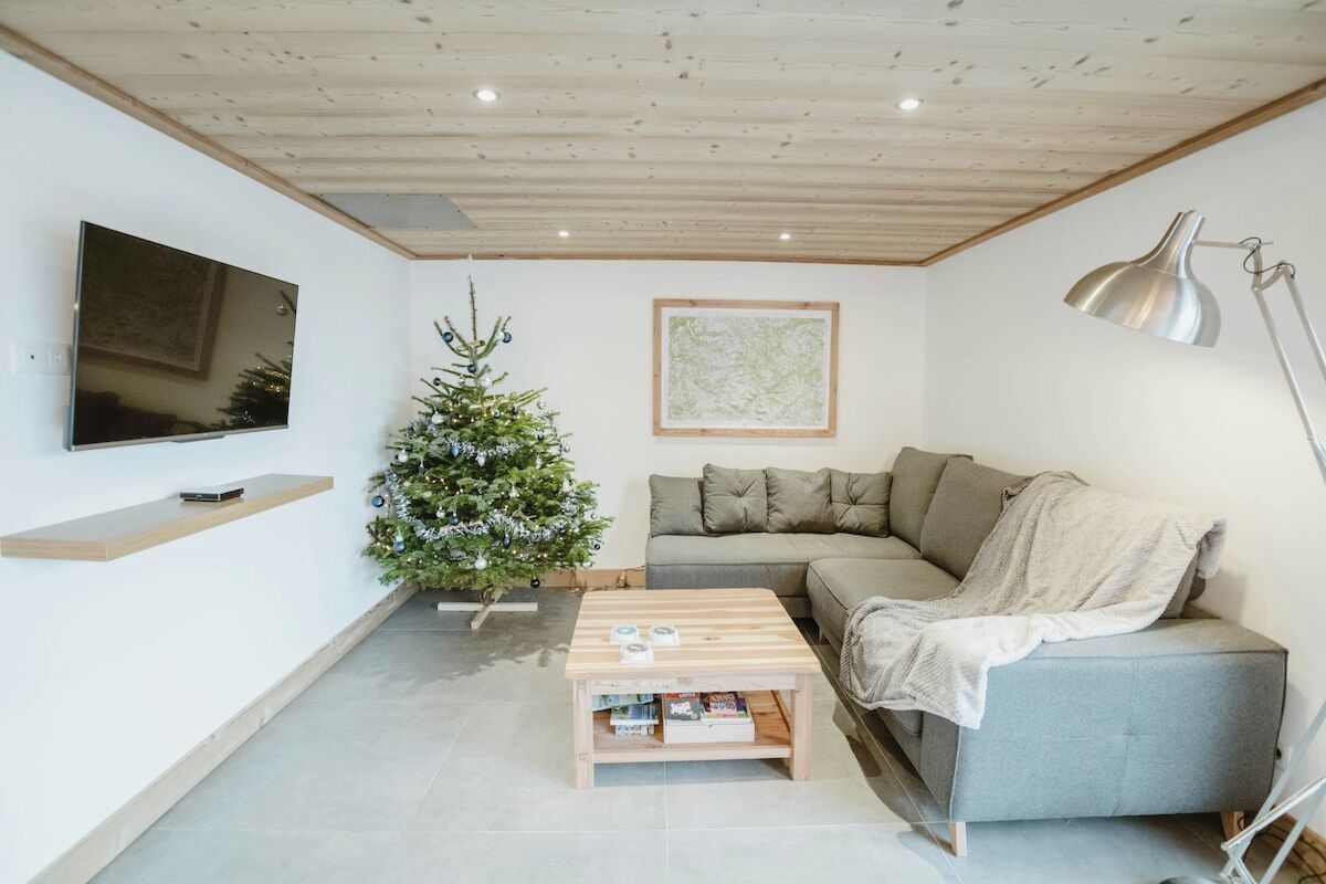 Ein Wohnzimmer mit einem Flachbildfernseher, einem Weihnachtsbaum, einem grauen Sofa, einem Holztisch und einer Stehlampe.
