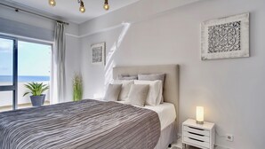 A bedroom with a large bed, a bedside table with a lamp, a potted plant, and a view of the sea. - Premium w terrace. 2 min 2 beach, 90 sqm, Algarve (Quarteira)