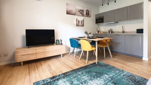Ein modernes Wohnzimmer mit Flachbildfernseher, einer Holzkommode, einem Esstisch mit bunten Stühlen sowie einer Küchenzeile mit grauen Schränken und einer Mikrowelle.