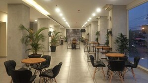 A modern indoor space with a long corridor lined with tables and chairs, potted plants, and a large window providing a view of the outside. - Arte Yogyakarta (Yogyakarta)