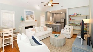 Ein modernes Wohnzimmer mit einem weißen Sofa, einem Couchtisch und einem Kamin. Im Hintergrund ist eine Küche mit Kühlschrank und Mikrowelle zu sehen.