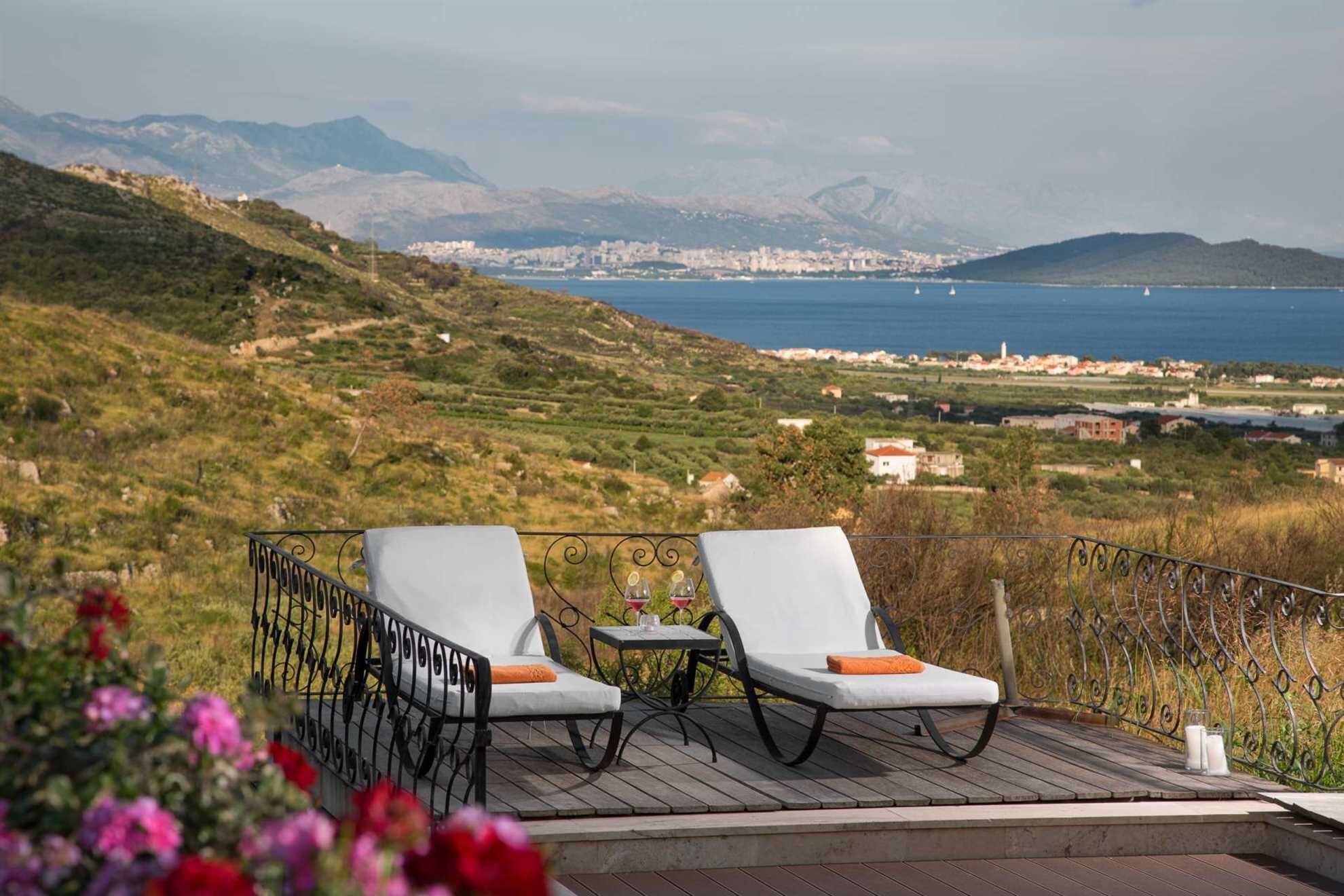 A terrace with two white lounge chairs and a small table, overlooking a coastal landscape with hills and a distant city.