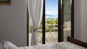 A bedroom with a bed, a window with white curtains, and a view of an outdoor patio with a table and chairs.
