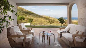 A cozy outdoor seating area with wicker furniture and a glass table, overlooking a scenic landscape with hills and a view of the sea.