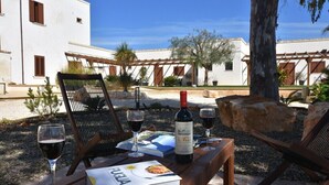 A wooden table with a bottle of wine, glasses, a menu, and a book in a sunny outdoor setting with a white building and trees in the background.