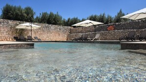 A clear pool with lounge chairs and umbrellas, surrounded by a stone wall and trees.
