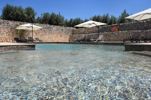 Piscina límpida com espreguiçadeiras e guarda-sóis, cercada por um muro de pedra e árvores.