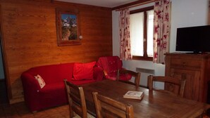 A cozy room with a wooden wall, a red sofa, a wooden dining table with chairs, a window with patterned curtains, and a television on a wooden cabinet.