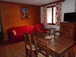 Um quarto aconchegante com parede de madeira, um sofá vermelho, uma mesa de jantar de madeira com cadeiras, uma janela com cortinas estampadas e uma televisão em um armário de madeira.