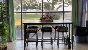 A dining area with a table and four chairs, a potted plant, and a view of a tree-lined field through large windows. - Sunshine & Beach Views at 2046 Beach Blvd (Biloxi)