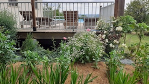 Um jardim com diversas plantas e flores, um deck de madeira com guarda-corpo, e vista para uma piscina ao fundo.