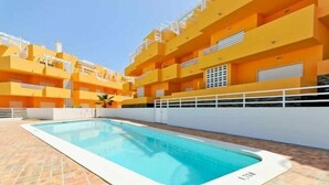 Une piscine entourée d’une terrasse carrelée, elle-même bordée d’immeubles jaunes à plusieurs étages, dotés de balcons.
