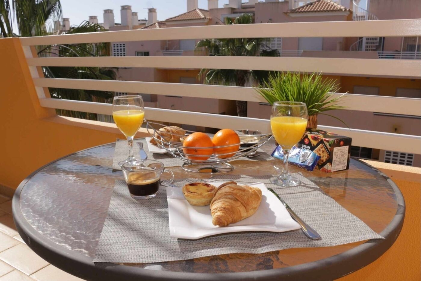 Ein Frühstückstisch mit Croissants, Orangensaft und Kaffee auf einem Balkon mit Blick auf Gebäude und Palmen.