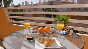 Une table de petit-déjeuner avec des croissants, du jus d’orange et du café, placée sur un balcon donnant sur des immeubles et des palmiers.