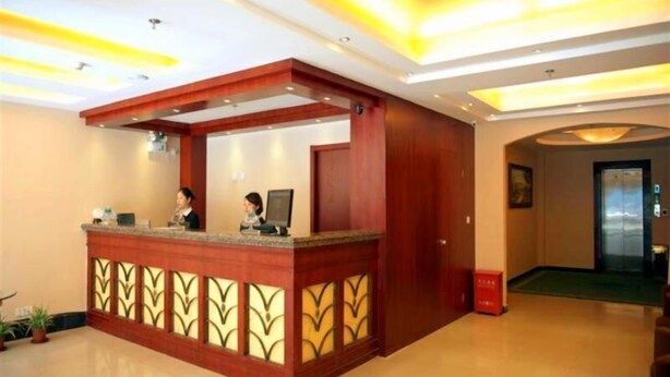 A hotel reception area with a wooden reception desk, two receptionists, and a green carpeted hallway.