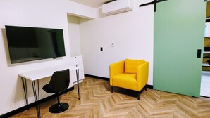 Habitación con un televisor de pantalla plana grande, un escritorio blanco con una silla negra, un sillón amarillo y una puerta corredera verde.