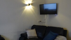 Un salon avec une table basse en bois, un canapé noir agrémenté de coussins bleus, un téléviseur à écran plat fixé au mur et un luminaire fixé au mur.