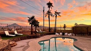 A swimming pool with lounge chairs and palm trees, surrounded by a stone wall and a sunset view. - Trendy Getaway + Pool, Spa, Game Room, Views (Lake Havasu City)