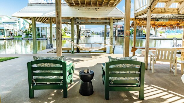 Eine Holzterrasse mit einer Hängematte, grünen Stühlen und einem Tisch an einem Gewässer.