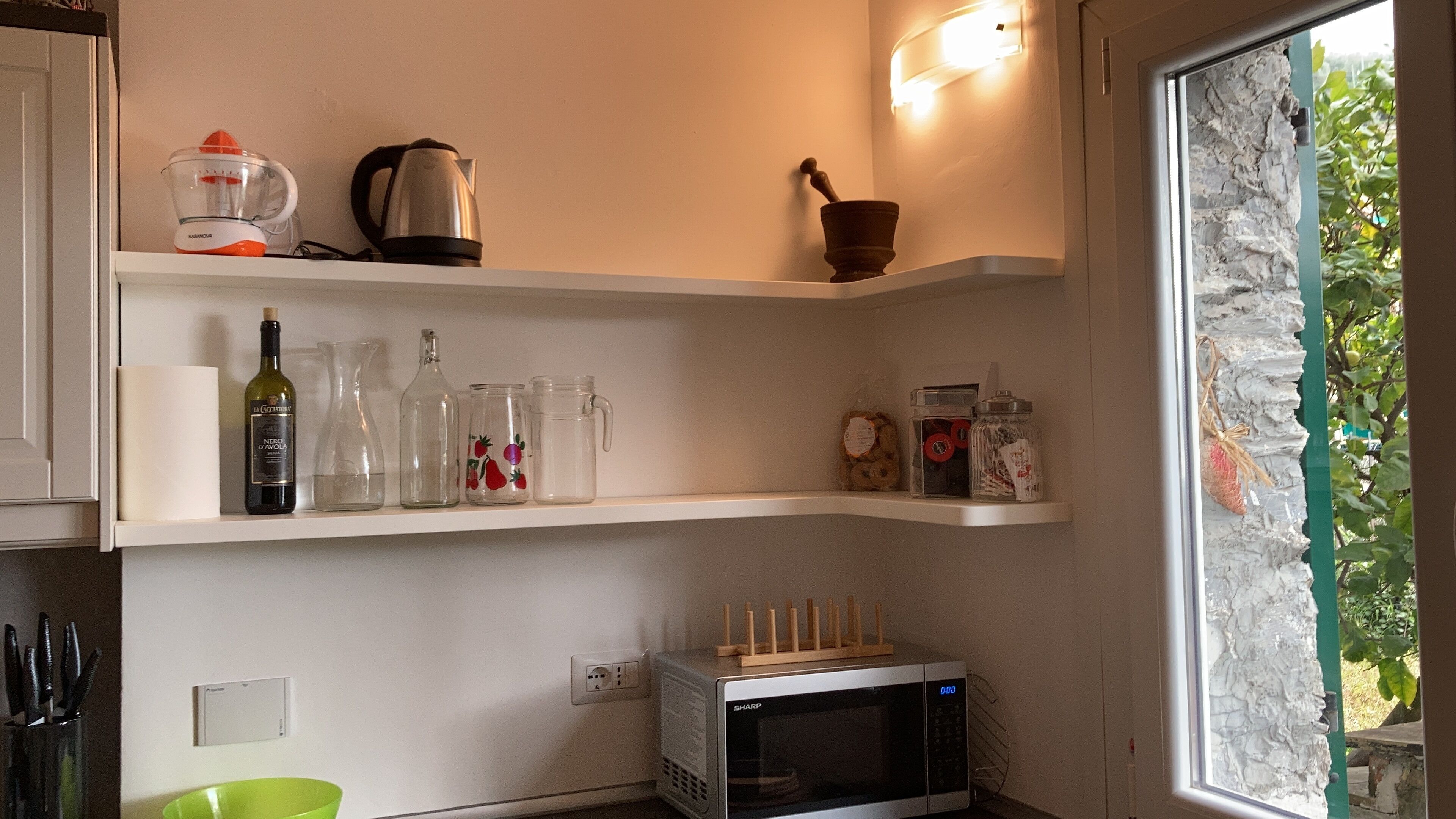 A kitchen with a microwave, shelves with kitchenware, and a window with a view of greenery.