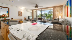 A modern kitchen with a central island, a dining area with a TV, and a balcony with a view of palm trees. - Island Surf Resort, breathtaking ocean view, in the heart of the action (Kihei)