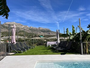 Ein Poolbereich mit Liegestühlen und Sonnenschirmen, eine Rasenfläche und eine Bergkulisse im Hintergrund.