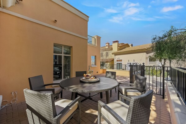 An outdoor patio with a round table and wicker chairs, a small table with a fruit bowl, and a view of neighboring buildings. - The Ideal Townhouse Getaway (Henderson)